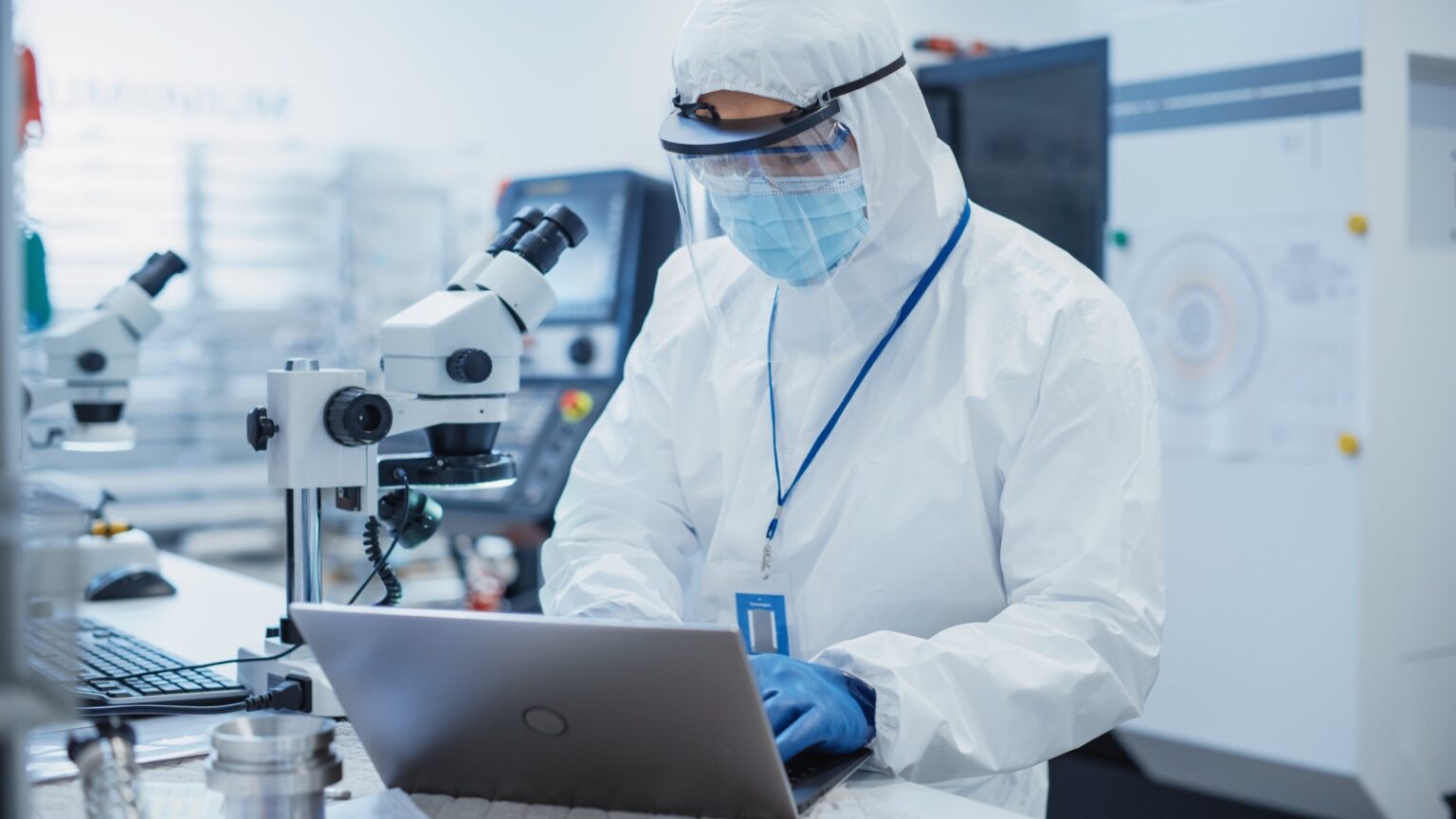 Scientist in protective gear working in a biotech cleanroom environment with microscopes and a laptop, representing advanced life sciences research and laboratory innovation.