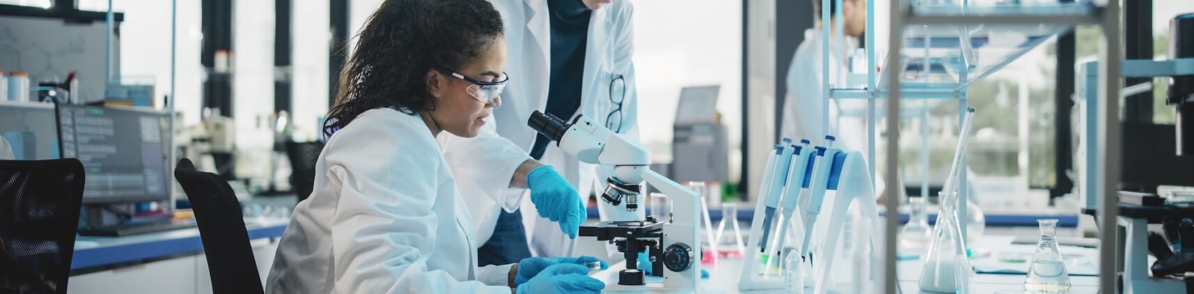 Two scientists working together in a laboratory, with a women seated looking in a microscope and a man standing adjusting microscope slides.