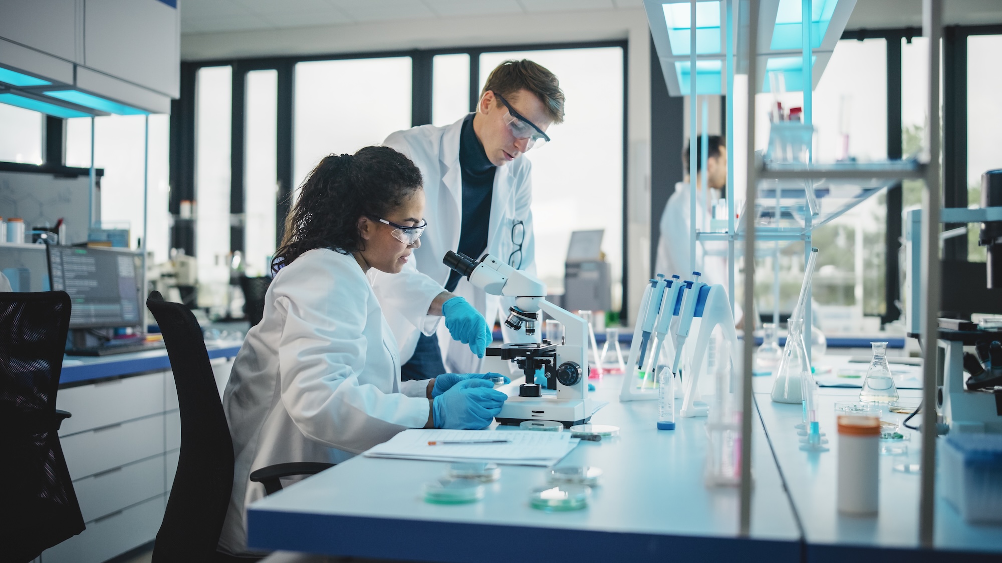 Two scientists working together in a laboratory, with a women seated looking in a microscope and a man standing adjusting microscope slides.