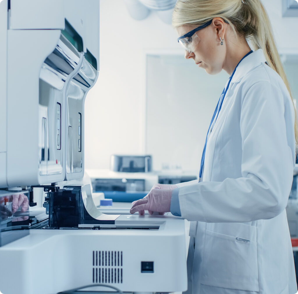 A laboratory technician in a white coat and safety glasses operates advanced analytical instrumentation in a modern life science laboratory environment.